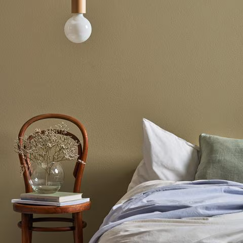 Bett vor einer grünen wand, daneben ein Holzstuhl mit Büchern und einer Vase und darüber eine Leuchte